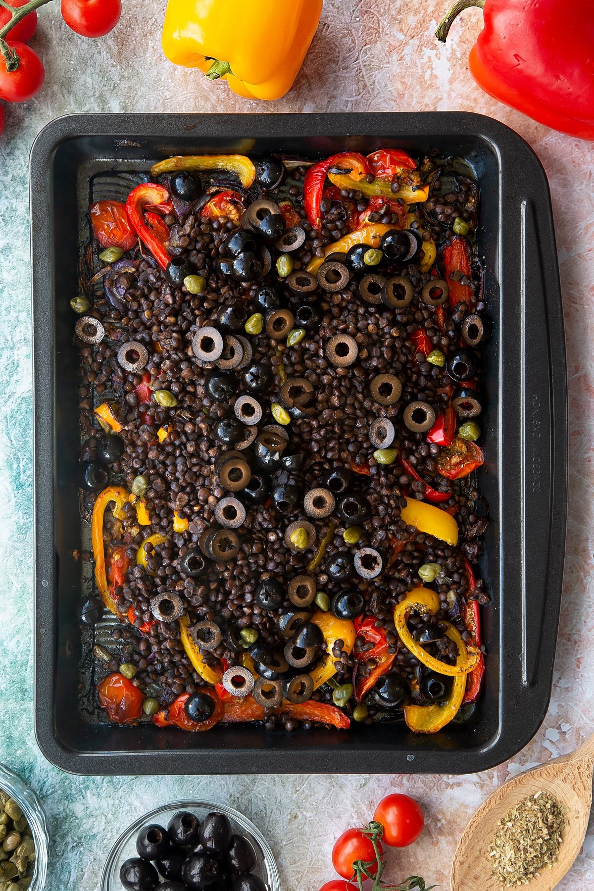 Roasted cherry tomatoes, peppers and onions in an oven tray, topped with cooked puy lentils, olives and capers. Ingredients to make vegan lentil salad surround the tray.