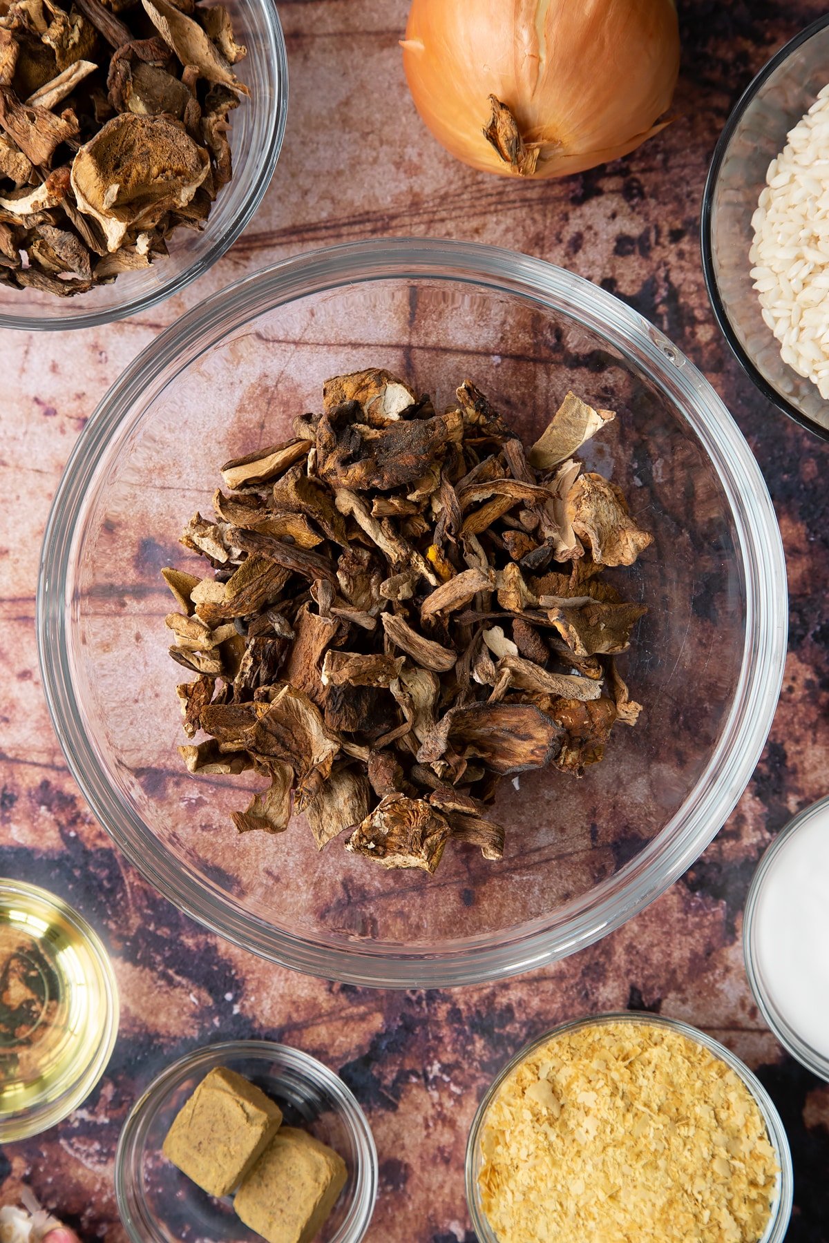 Dried porcini mushrooms in a glass bowl. Ingredients to make vegan mushroom risotto surround the bowl.