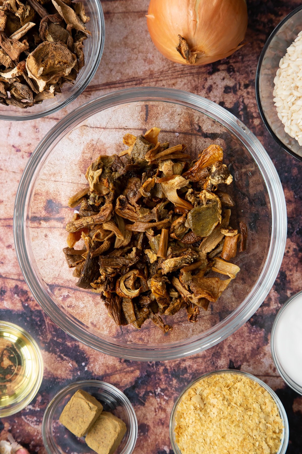 Soaked porcini mushrooms in a glass bowl. Ingredients to make vegan mushroom risotto surround the bowl.