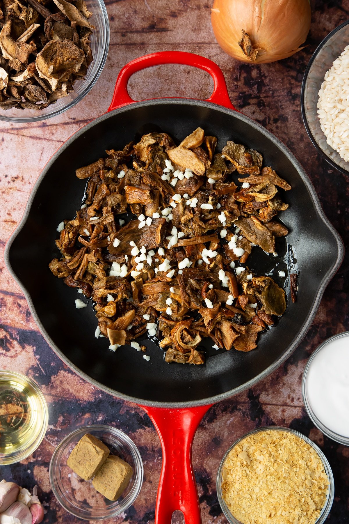 Soaked porcini mushrooms, oil and garlic in a pan. Ingredients to make vegan mushroom risotto surround the pan.