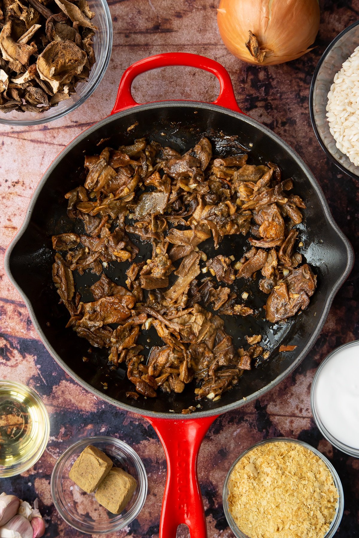 Fried porcini mushrooms, oil and garlic in a pan. Ingredients to make vegan mushroom risotto surround the pan.