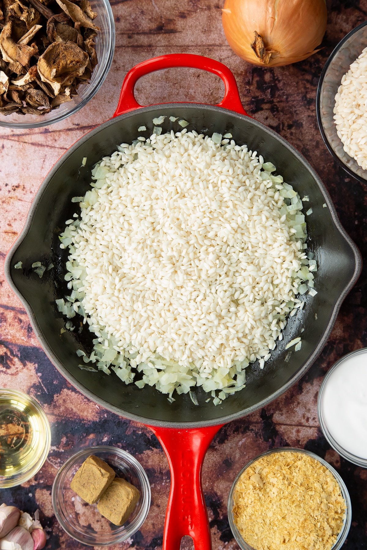 Lightly fried onions with arborio rice on top in a pan. Ingredients to make vegan mushroom risotto surround the pan.