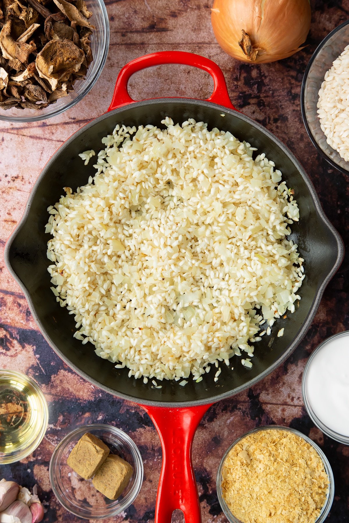 Lightly fried onions and arborio rice in a pan. Ingredients to make vegan mushroom risotto surround the pan.