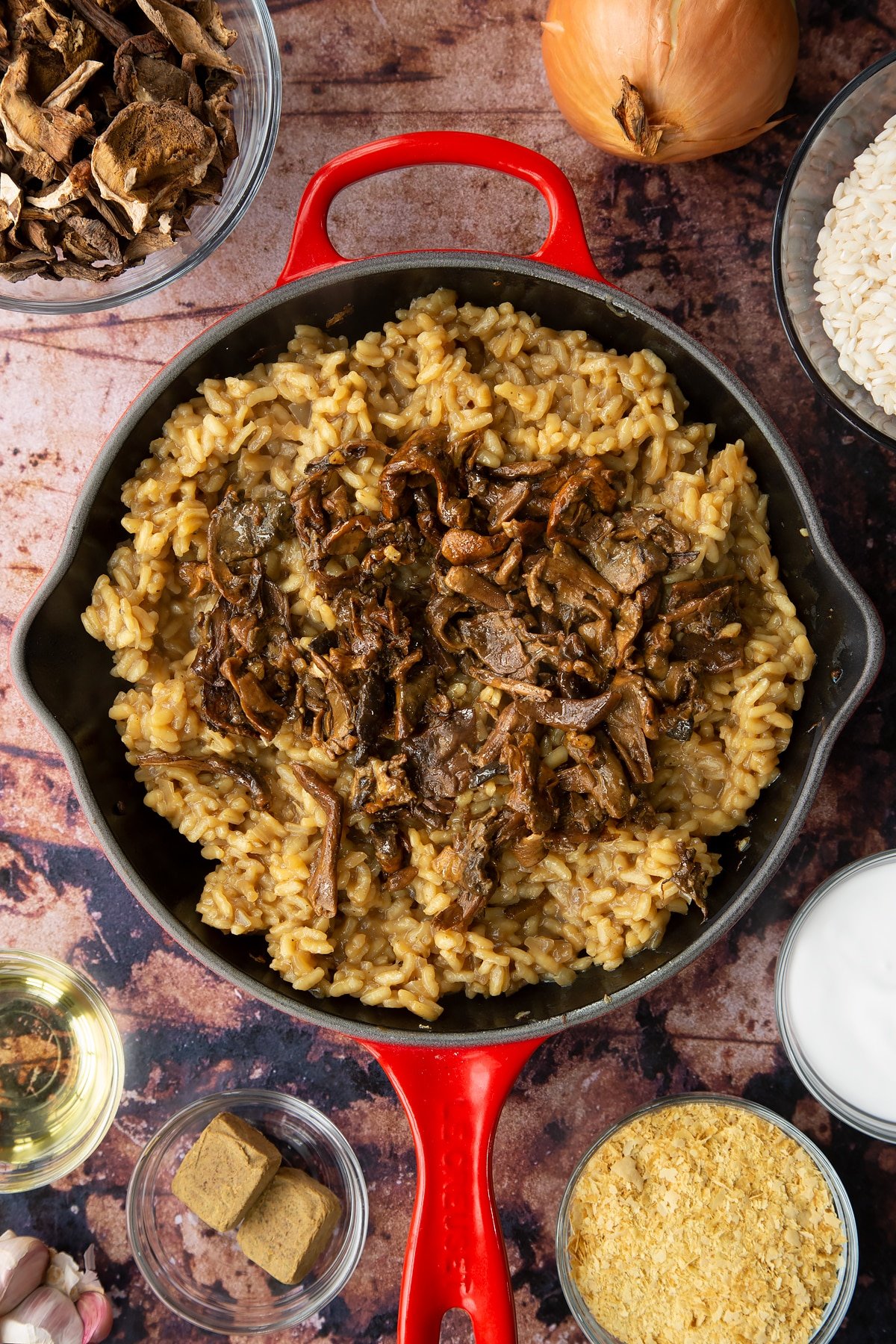 Mushroom risotto in a pan with fried porcini mushrooms on top. Ingredients to make vegan mushroom risotto surround the pan.