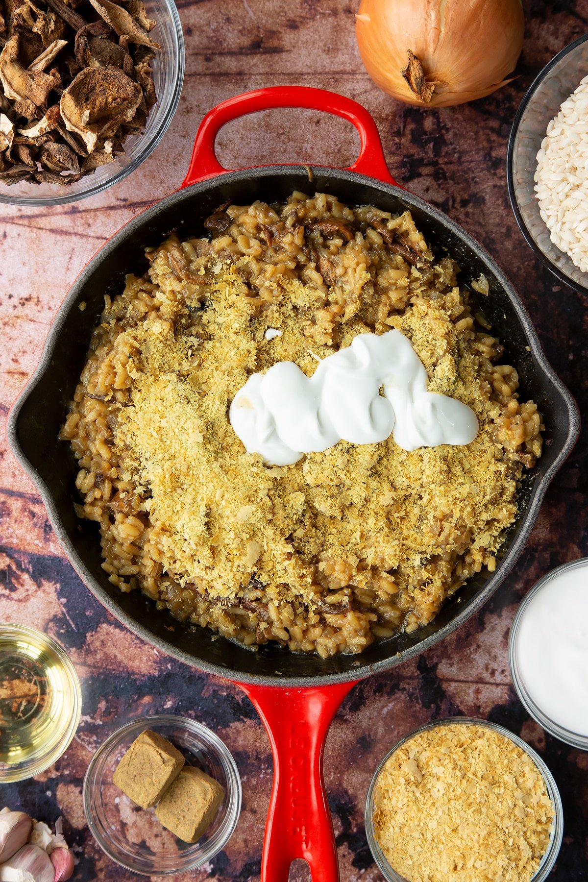 Mushroom risotto with fried porcini mushrooms in a pan with nutritional yeast and coconut cream on top. Ingredients to make vegan mushroom risotto surround the pan.