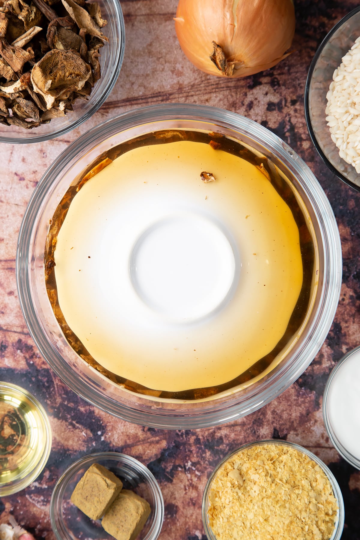 Dried porcini mushrooms covered with water and a small saucer in a glass bowl. Ingredients to make vegan mushroom risotto surround the bowl.