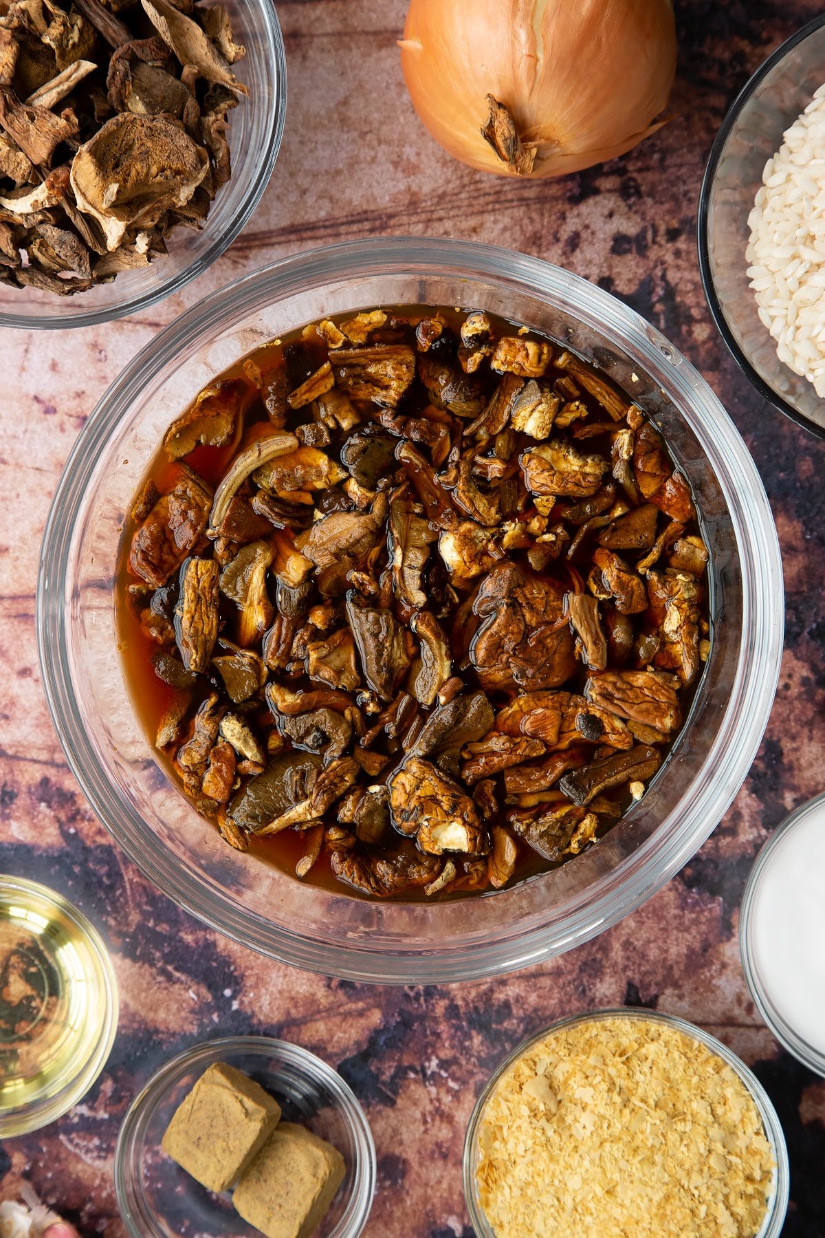 Porcini mushrooms soaked in water in a glass bowl. Ingredients to make vegan mushroom risotto surround the bowl.