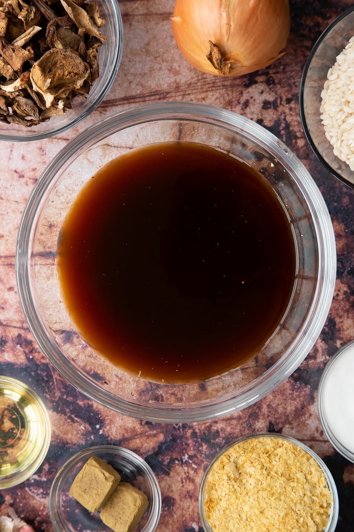 Mushroom stock from soaking porcini mushrooms in a glass bowl. Ingredients to make vegan mushroom risotto surround the bowl.