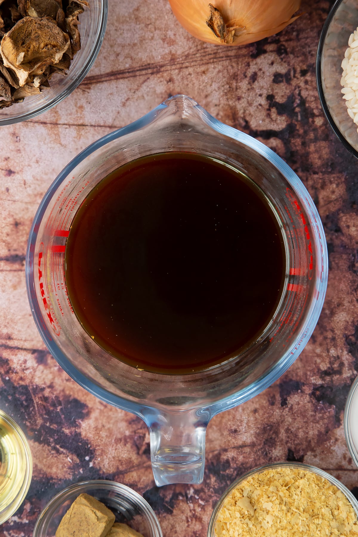 Mushroom stock from soaking porcini mushrooms in a glass jug. Ingredients to make vegan mushroom risotto surround the jug.