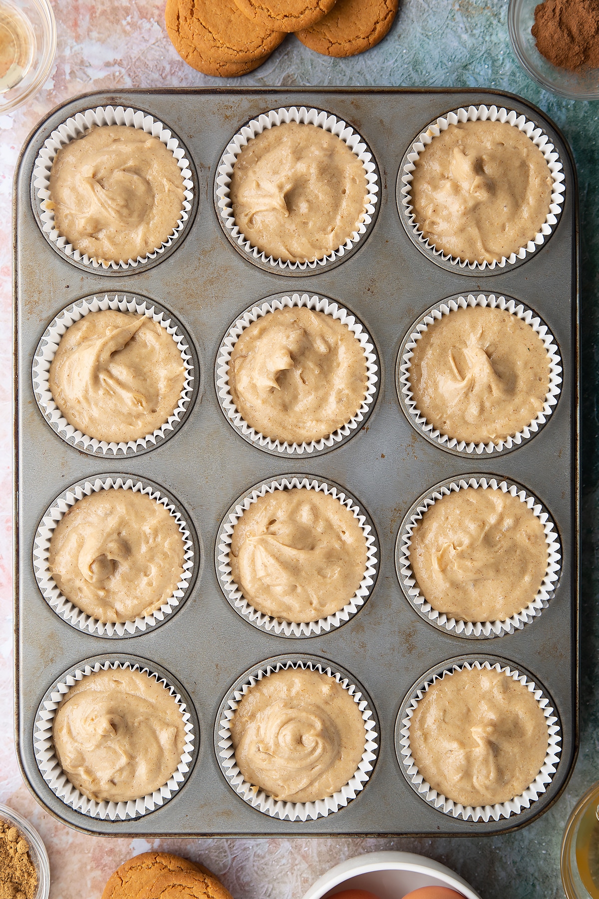 Muffin tin lined with cupcake cases filled with ginger cake batter. Ingredients to make ginger cupcakes surround the tin.