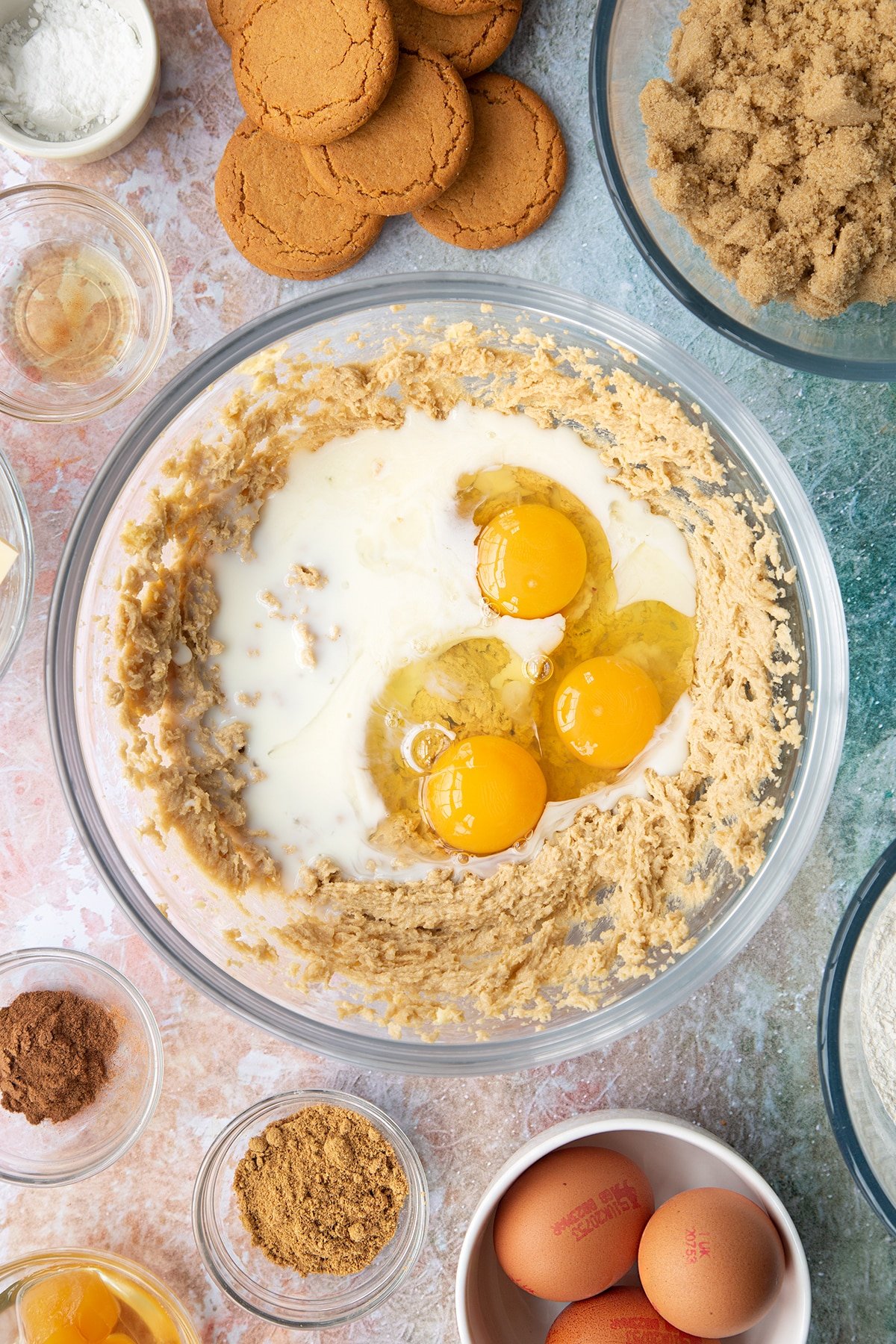 Butter and soft light brown sugar whisked together in a mixing bowl with milk, vanilla and eggs on top. Ingredients to make ginger cupcakes surround the bowl.