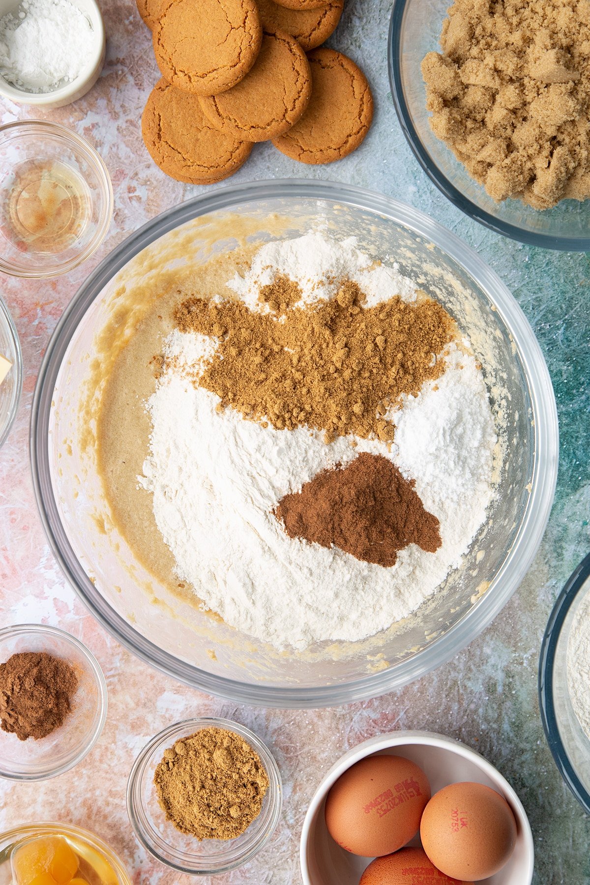 Butter, soft light brown sugar, milk, eggs and vanilla whisked together in a mixing bowl with flour, cinnamon and ginger on top. Ingredients to make ginger cupcakes surround the bowl.