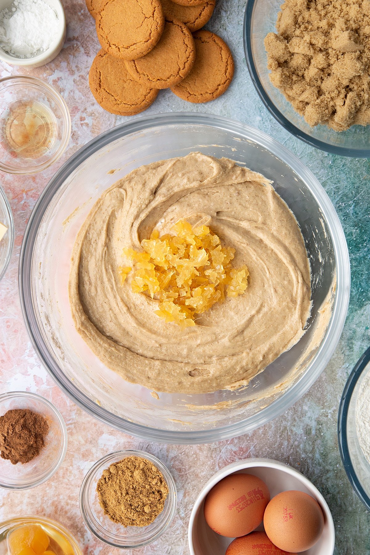 Ginger cake batter in a mixing bowl with chopped stem ginger on top. Ingredients to make ginger cupcakes surround the bowl.