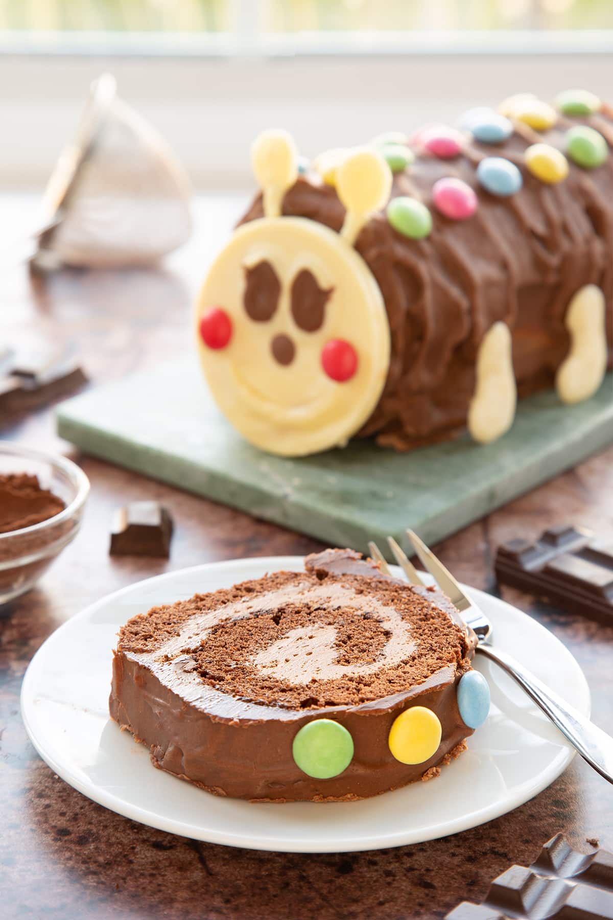 A slice of caterpillar cake on a small white plate with a fork. There is a caterpillar cake in the background.