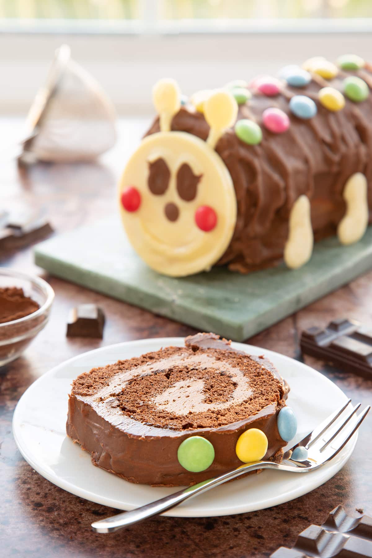 A slice of caterpillar cake on a small white plate with a fork beside it. There is a caterpillar cake in the background.