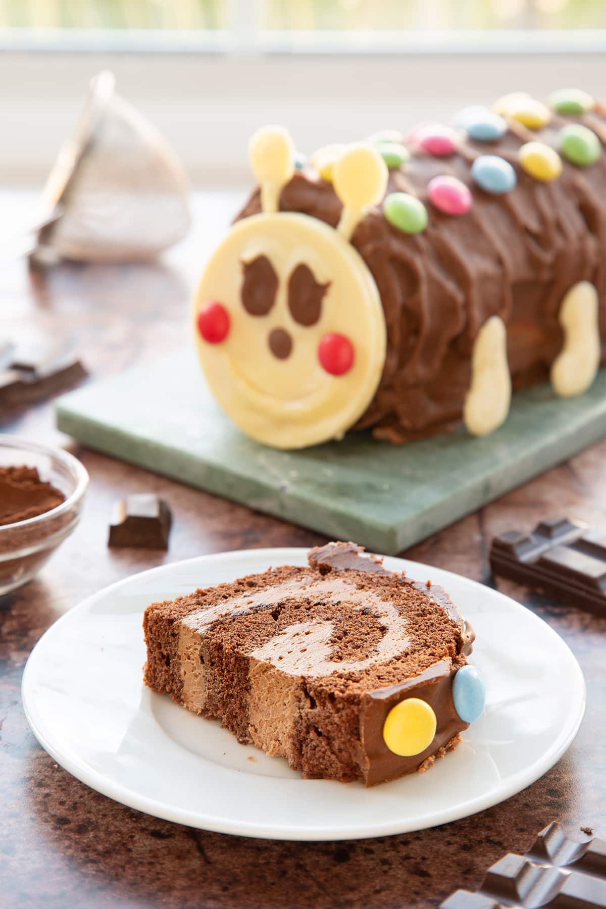 A slice of caterpillar cake on a small white plate. Part has been cut away. There is a caterpillar cake in the background.