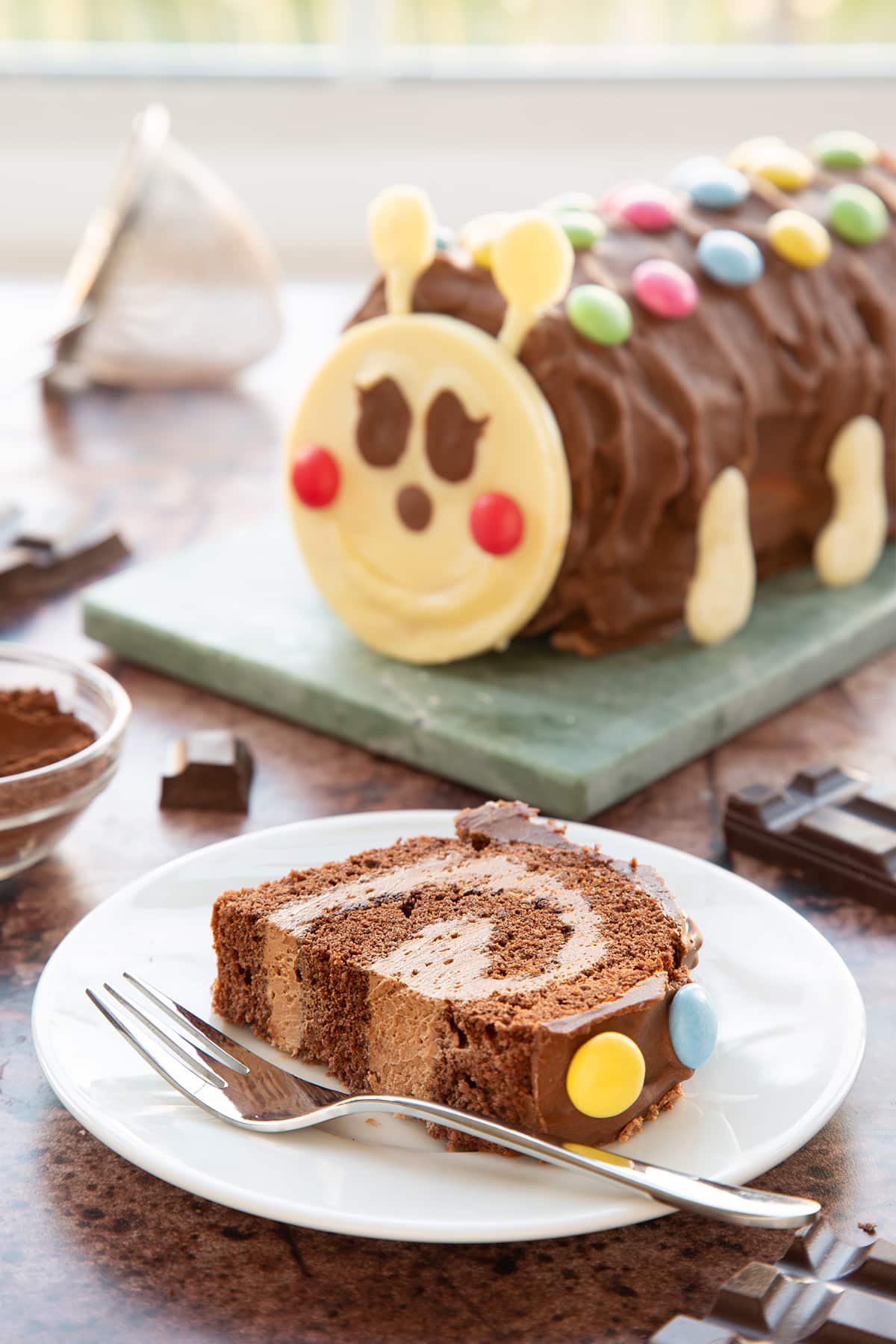 A slice of caterpillar cake on a small white plate with a fork. Part has been cut away. There is a caterpillar cake in the background.