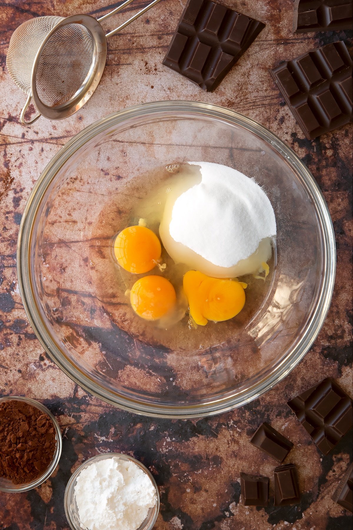 Eggs and caster sugar in a glass mixing bowl. Ingredients for the caterpillar cake recipe surround the bowl.