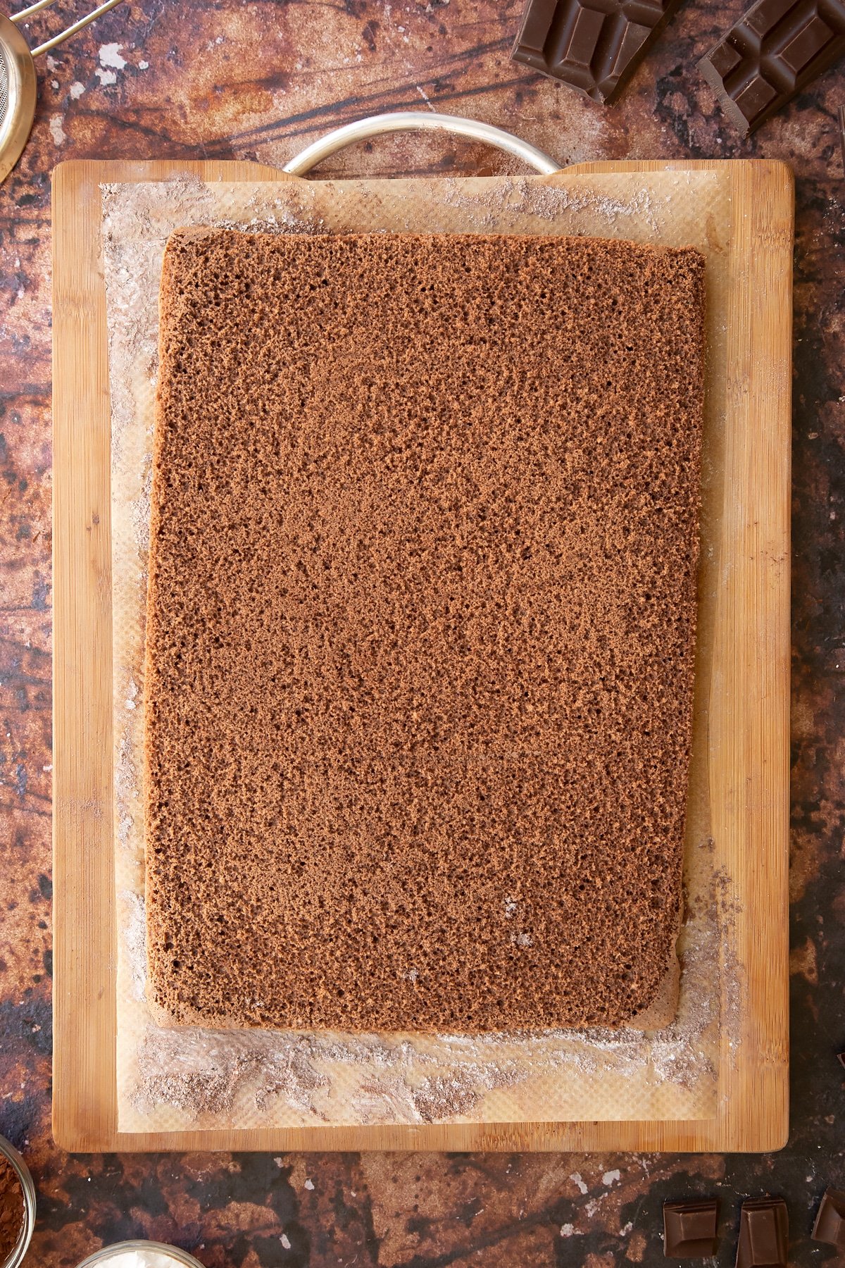 A rectangle of chocolate sponge on a wooden board and baking paper dusted with cocoa and icing sugar.