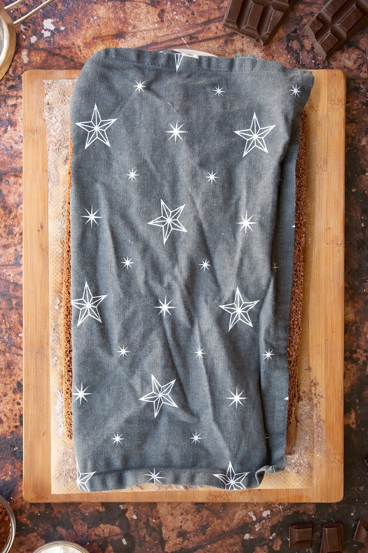 A rectangle of chocolate sponge with a tea towel one top on a wooden board and baking paper dusted with cocoa and icing sugar.