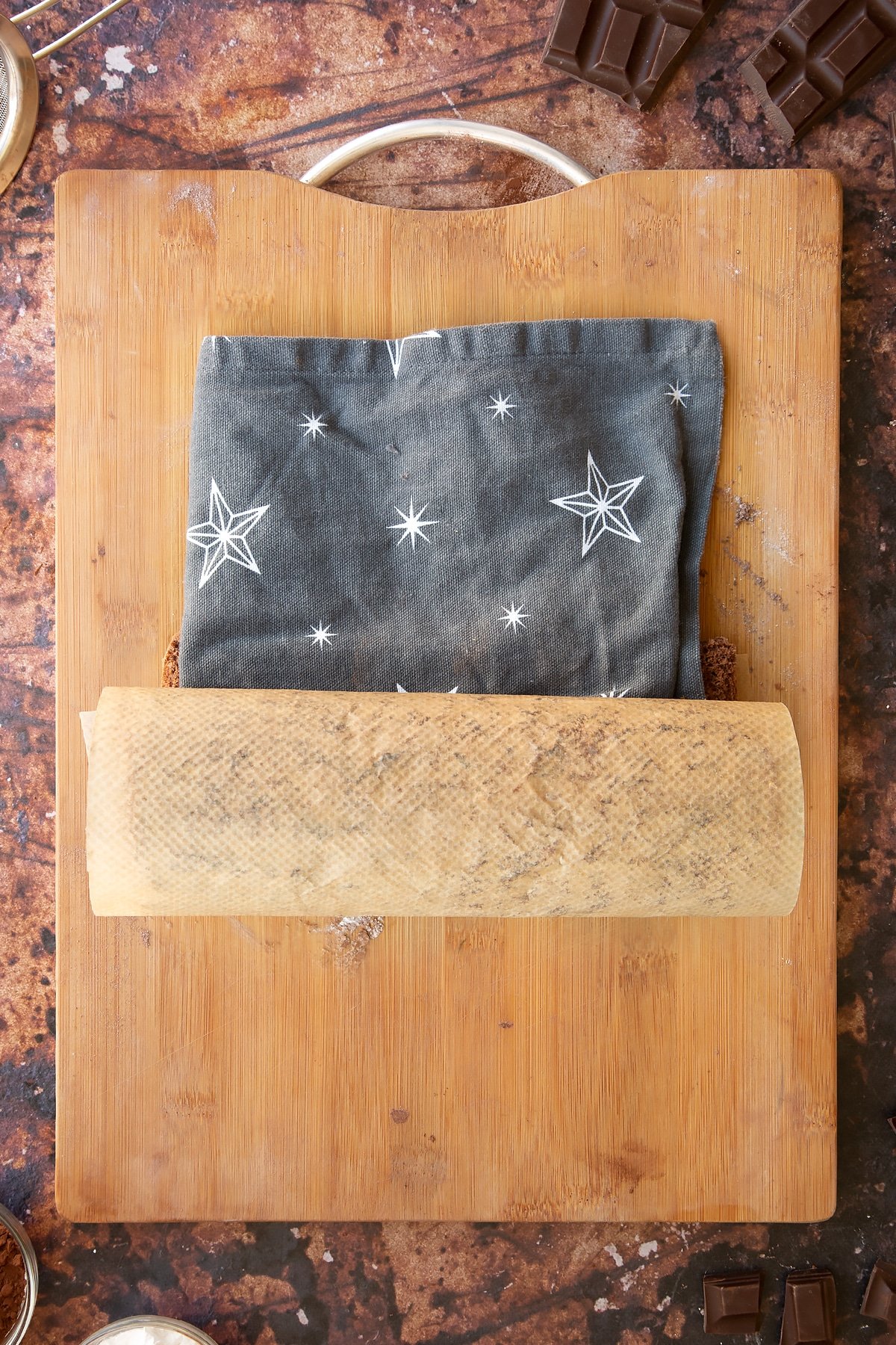 A chocolate sponge rolled with a tea towel on a wooden board and baking paper dusted with cocoa and icing sugar.