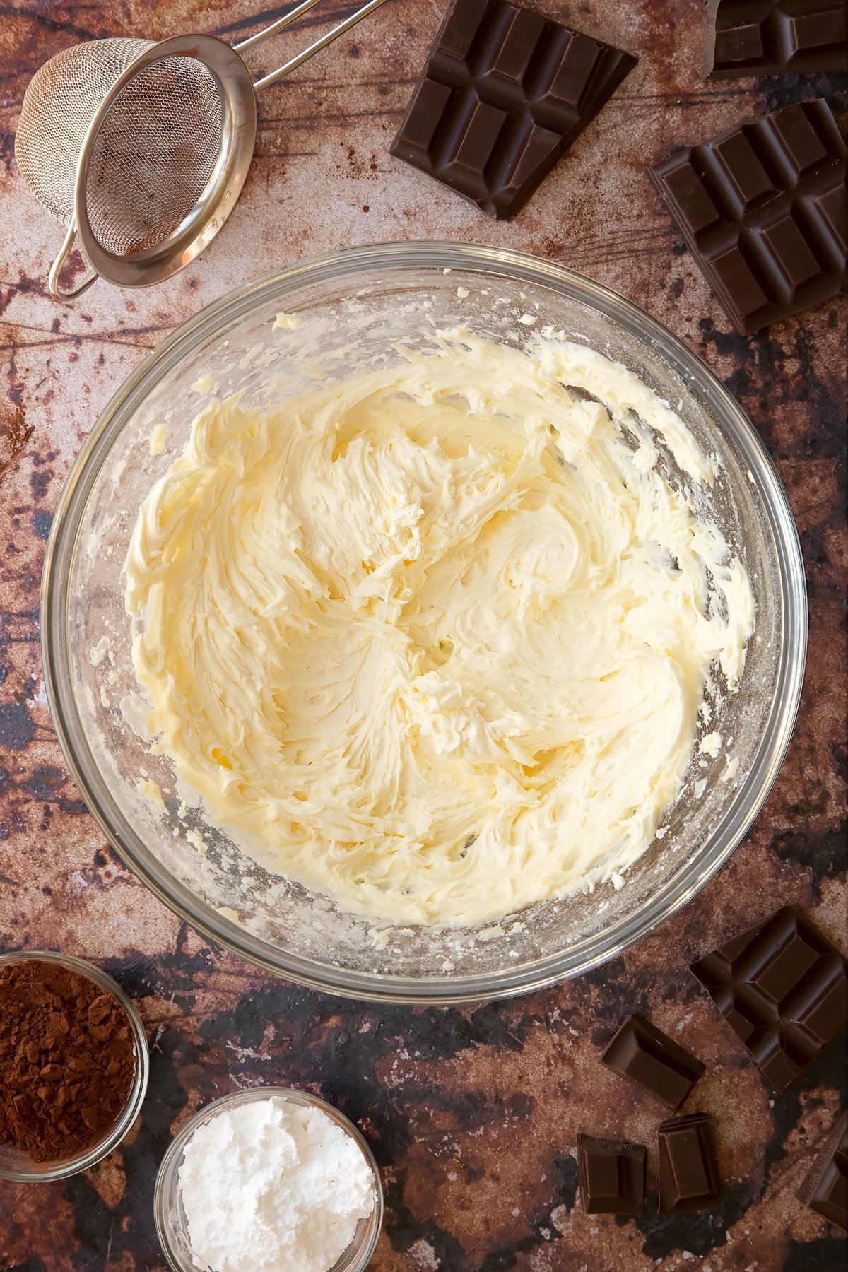 Butter, milk, vanilla and icing sugar whisked together in a glass mixing bowl. Ingredients for the caterpillar cake recipe surround the bowl.