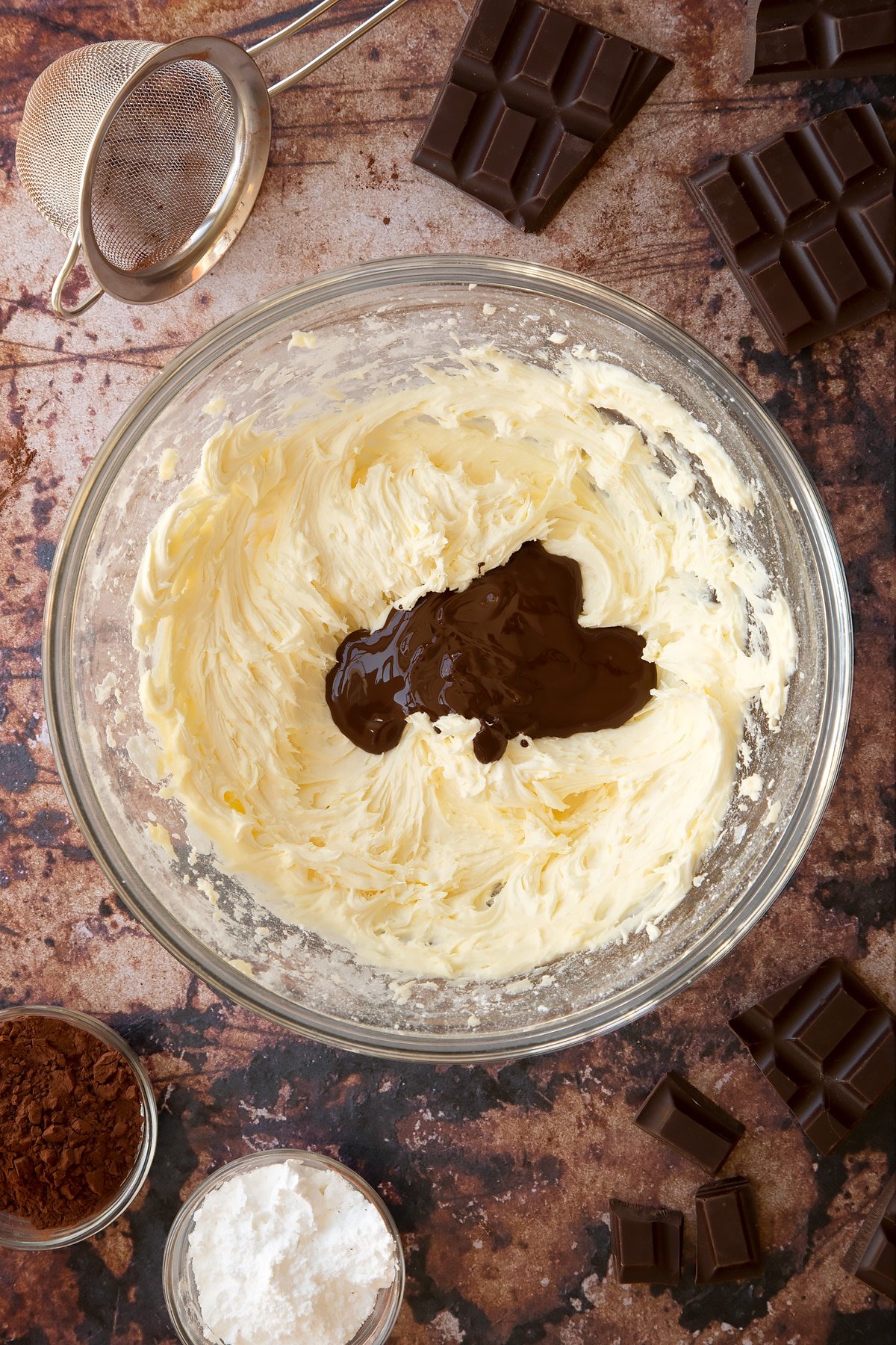 Butter, milk, vanilla and icing sugar whisked together in a glass mixing bowl with dark chocolate on top. Ingredients for the caterpillar cake recipe surround the bowl.