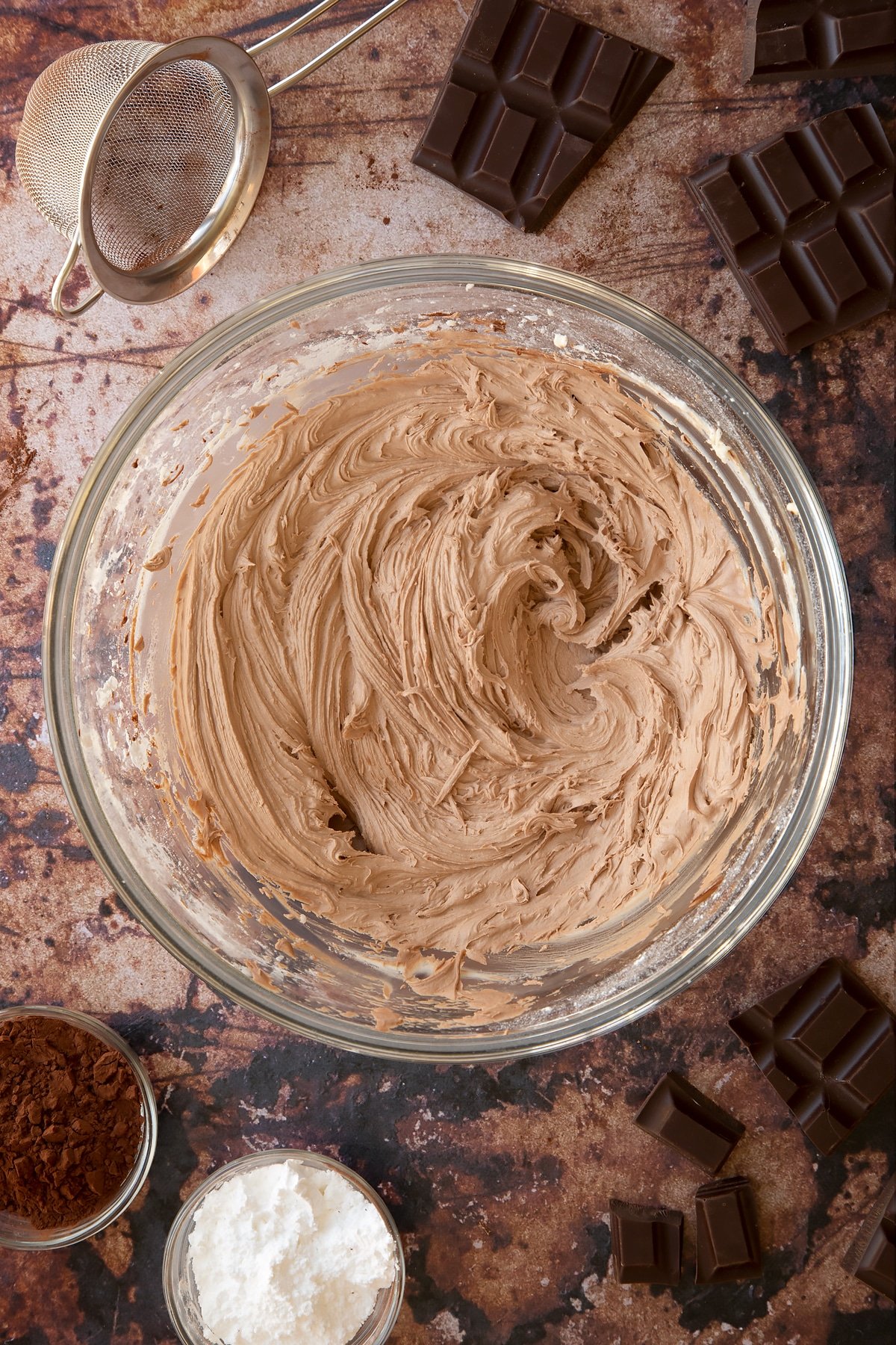 Chocolate buttercream in a glass mixing bowl. Ingredients for the caterpillar cake recipe surround the bowl.