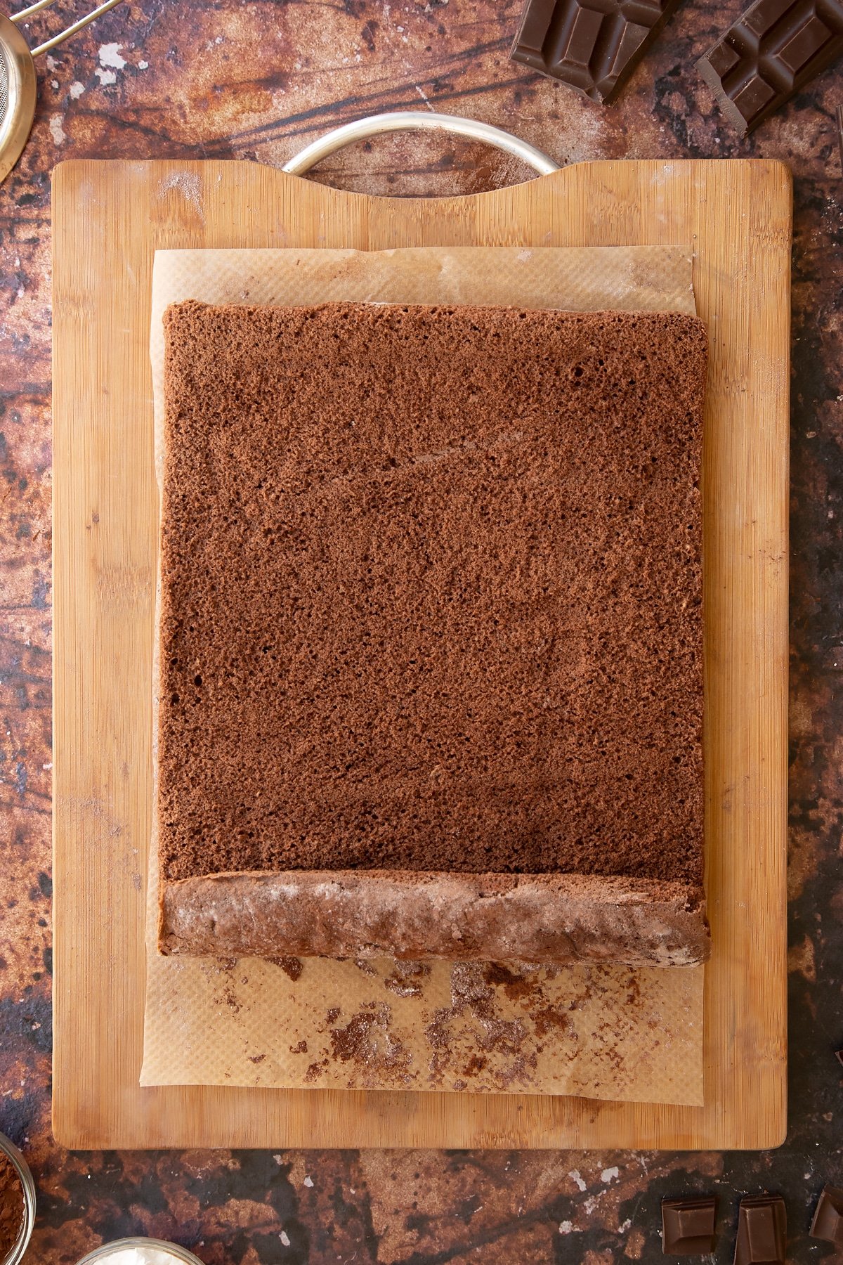 A chocolate sponge unrolled on a wooden board and baking paper dusted with cocoa and icing sugar. 