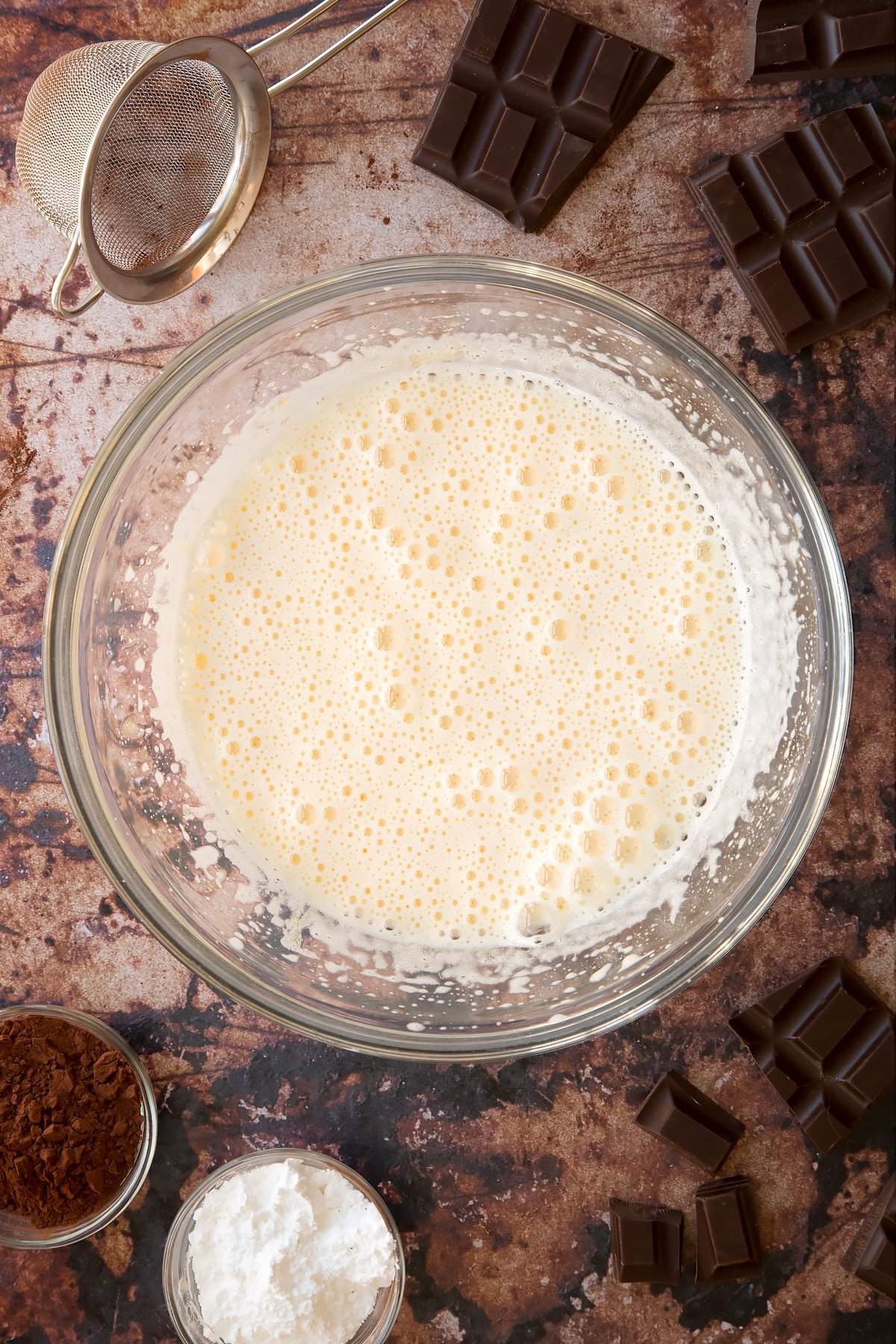 Eggs and caster sugar whisked together in a glass mixing bowl. Ingredients for the caterpillar cake recipe surround the bowl.