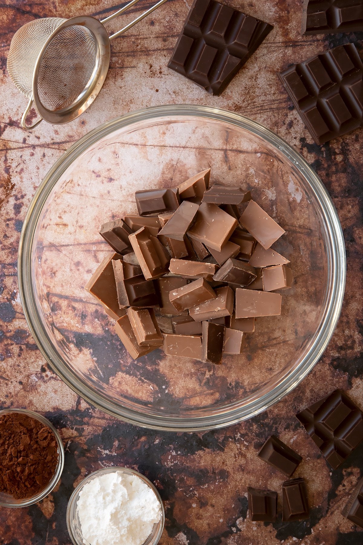 Milk and dark chocolate pieces in a glass mixing bowl. Ingredients for the caterpillar cake recipe surround the bowl.