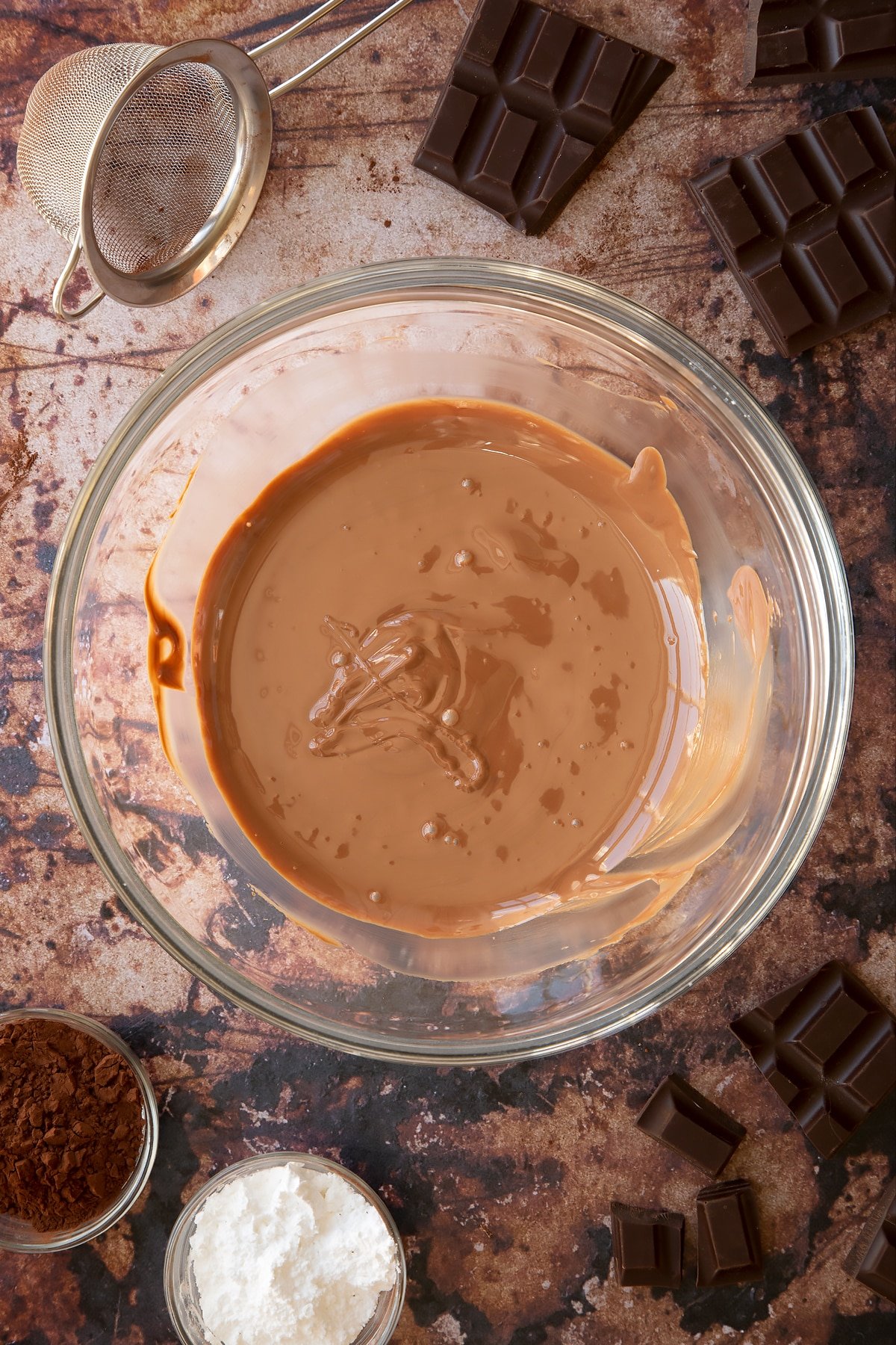 Melted chocolate in a glass mixing bowl. Ingredients for the caterpillar cake recipe surround the bowl.