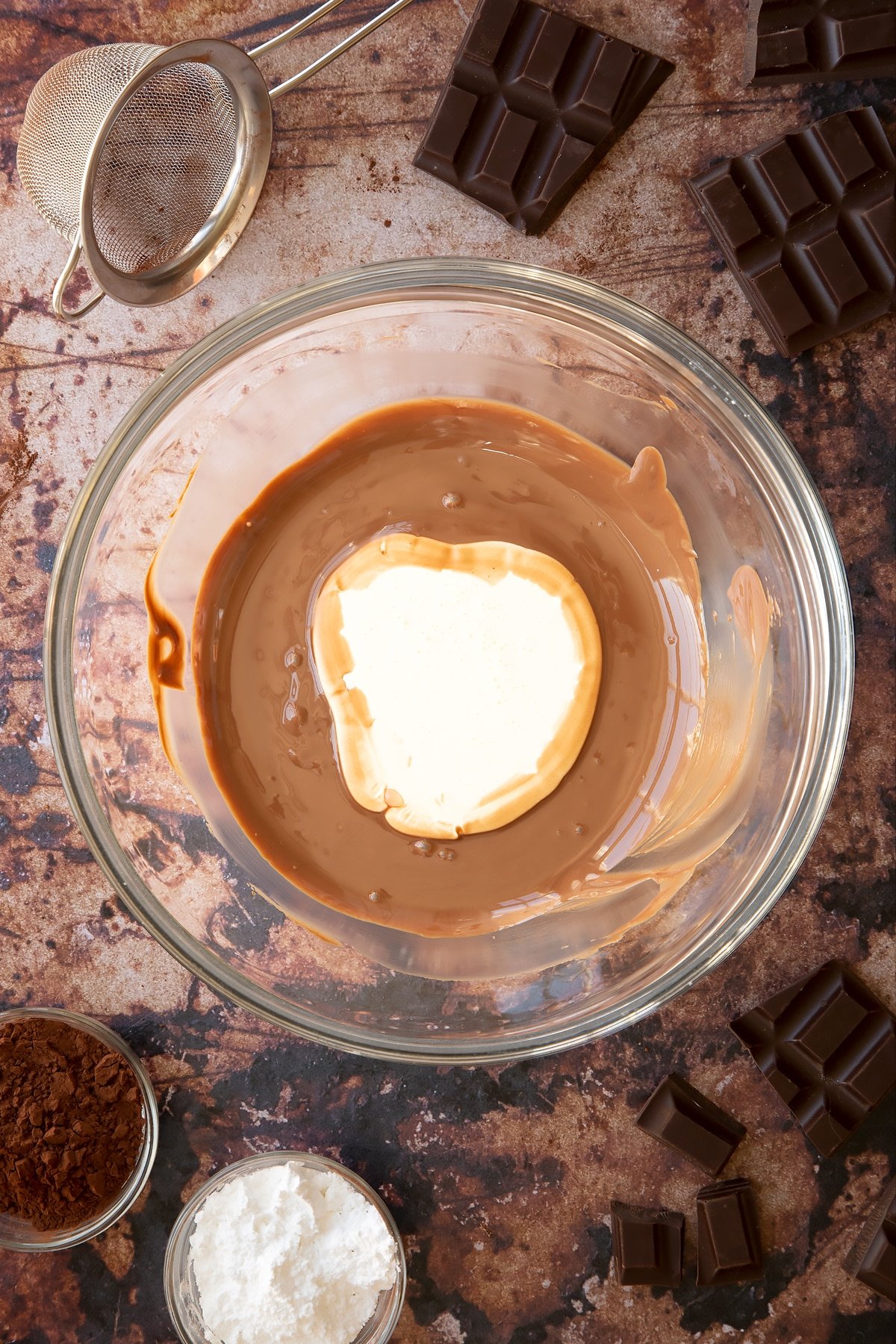 Melted chocolate in a glass mixing bowl with double cream on top. Ingredients for the caterpillar cake recipe surround the bowl.