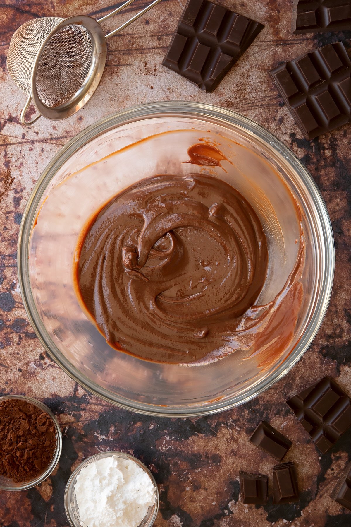 Chocolate ganache in a glass mixing bowl. Ingredients for the caterpillar cake recipe surround the bowl.