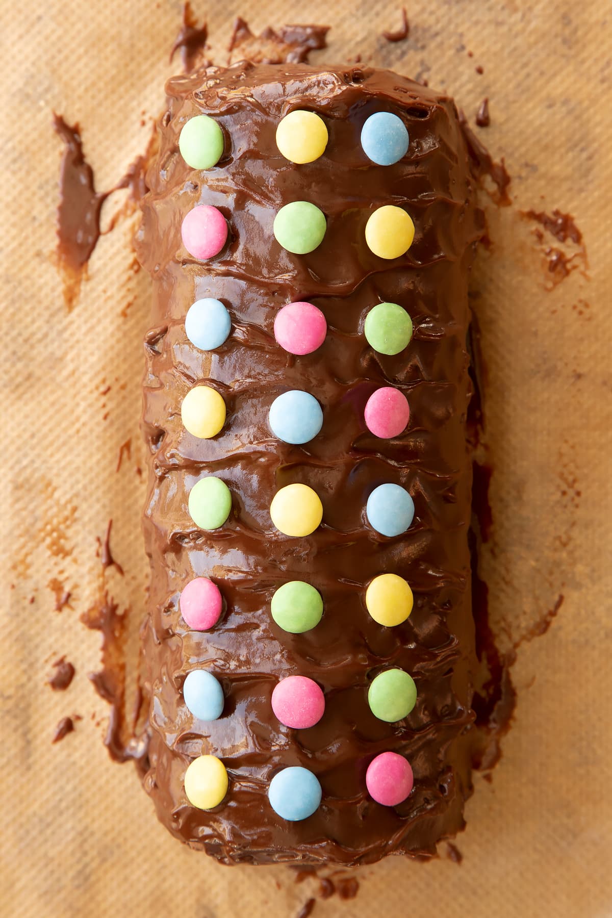 Chocolate Swiss roll covered with chocolate ganache and decorated with Smarties on baking paper.