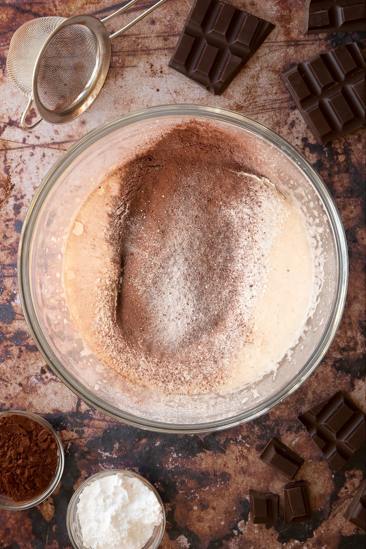Whisked eggs and caster sugar with cocoa and flour in a glass mixing bowl. Ingredients for the caterpillar cake recipe surround the bowl.