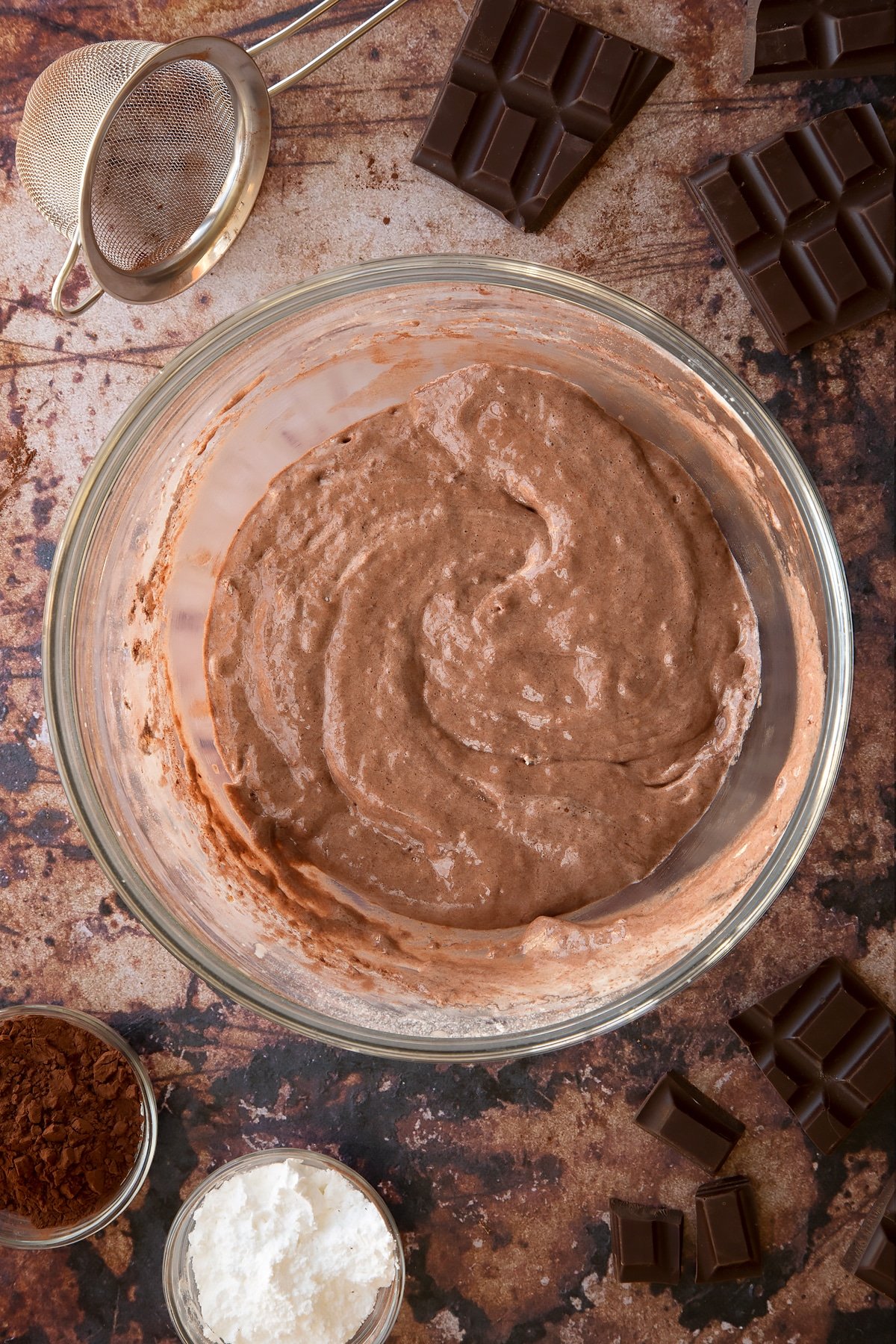 Chocolate sponge batter in a glass mixing bowl. Ingredients for the caterpillar cake recipe surround the bowl.