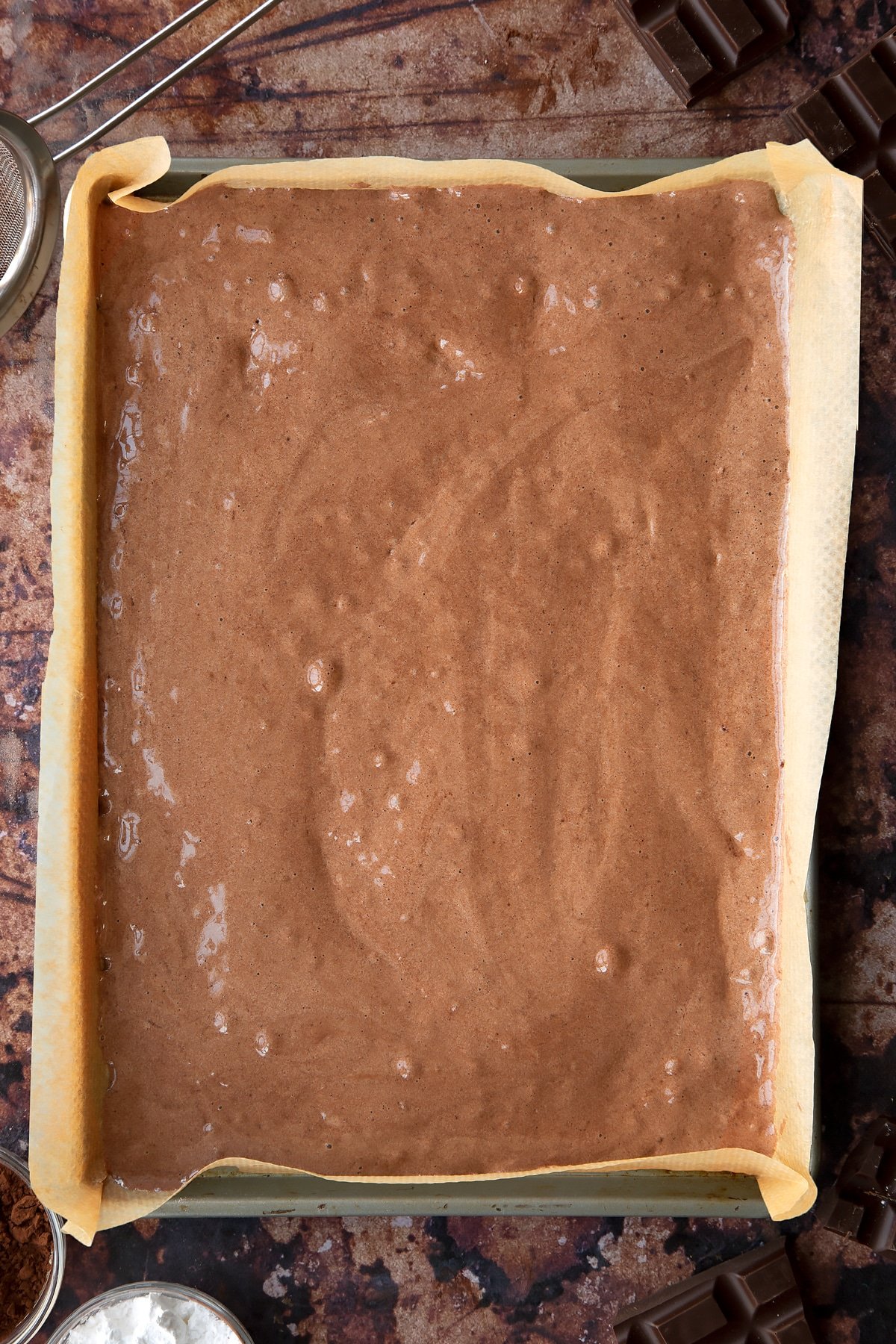 Chocolate sponge batter in a lined Swiss roll tray. Ingredients for the caterpillar cake recipe surround the tray.