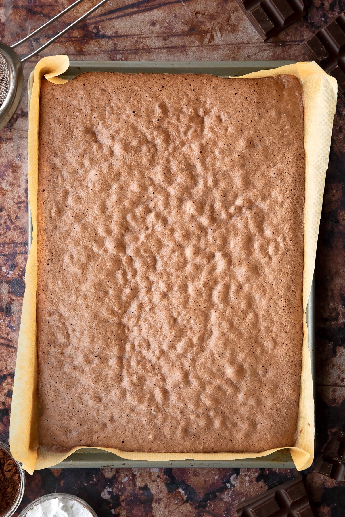 Cooked chocolate sponge in a lined Swiss roll tray. Ingredients for the caterpillar cake recipe surround the tray.