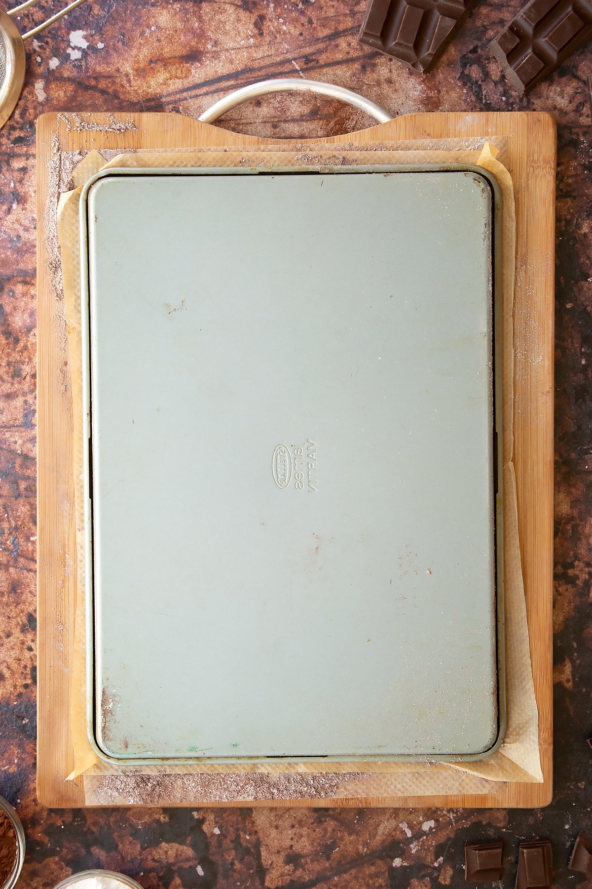 A wooden board and baking paper dusted with cocoa and icing sugar. A Swiss roll tin is upturned on the board.