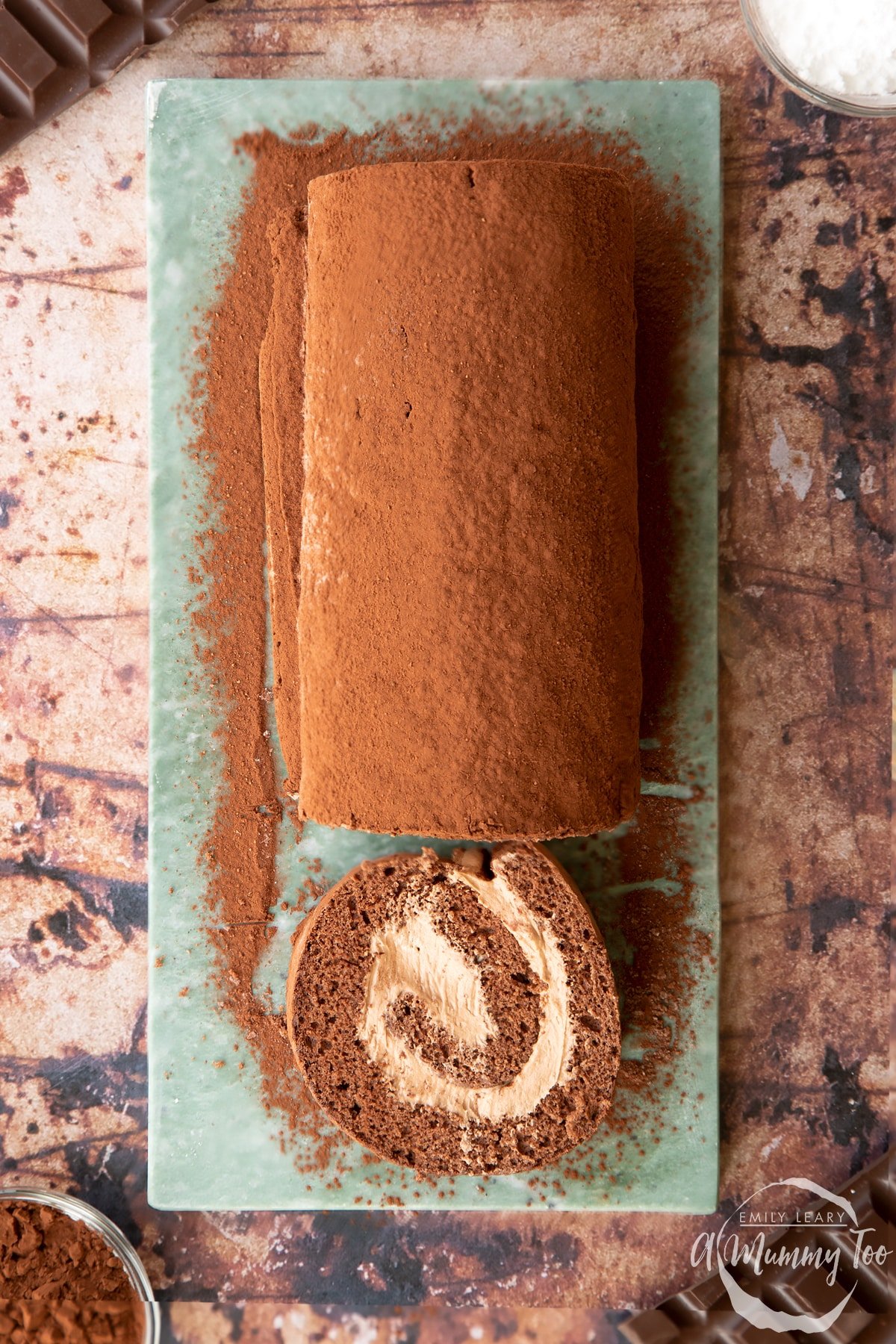 Chocolate Swiss roll dusted with cocoa on a grey marble board, shown from above. A slice has been cut.