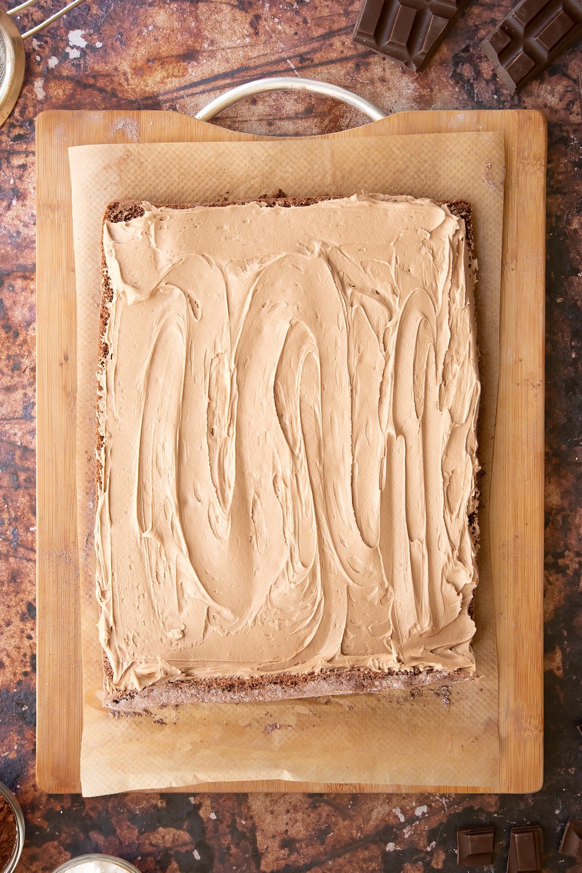 Wooden board with a chocolate Swiss roll sponge partially rolled up on it. The sponge is spread with chocolate buttercream.