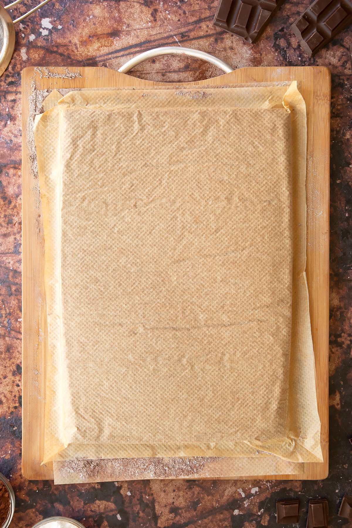 Wooden board with a chocolate Swiss roll sponge turned out onto it, covered with baking paper. 