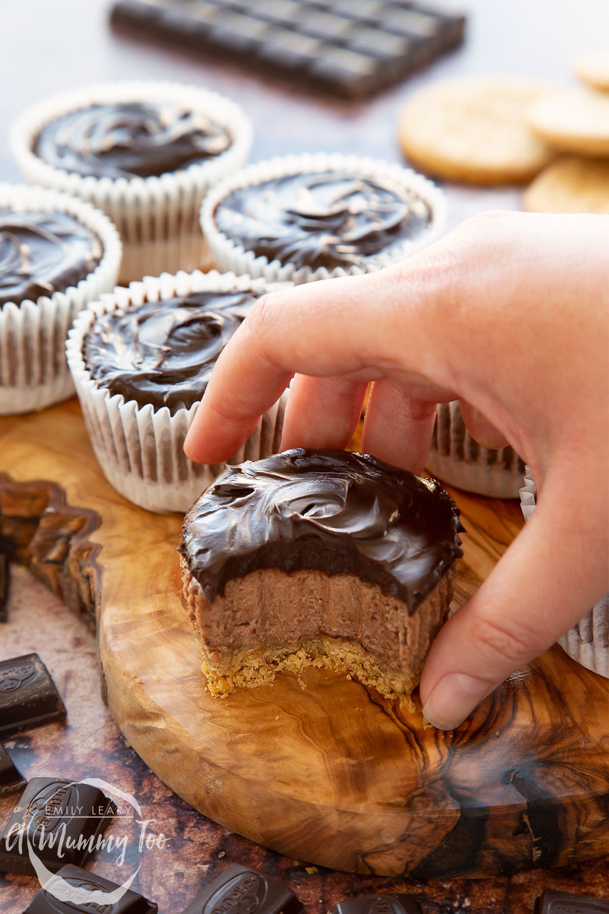 Chocolate cheesecake cupcakes in rows on an olive wood board. The one in the foreground has a bite out of it and a hand reaches for it.