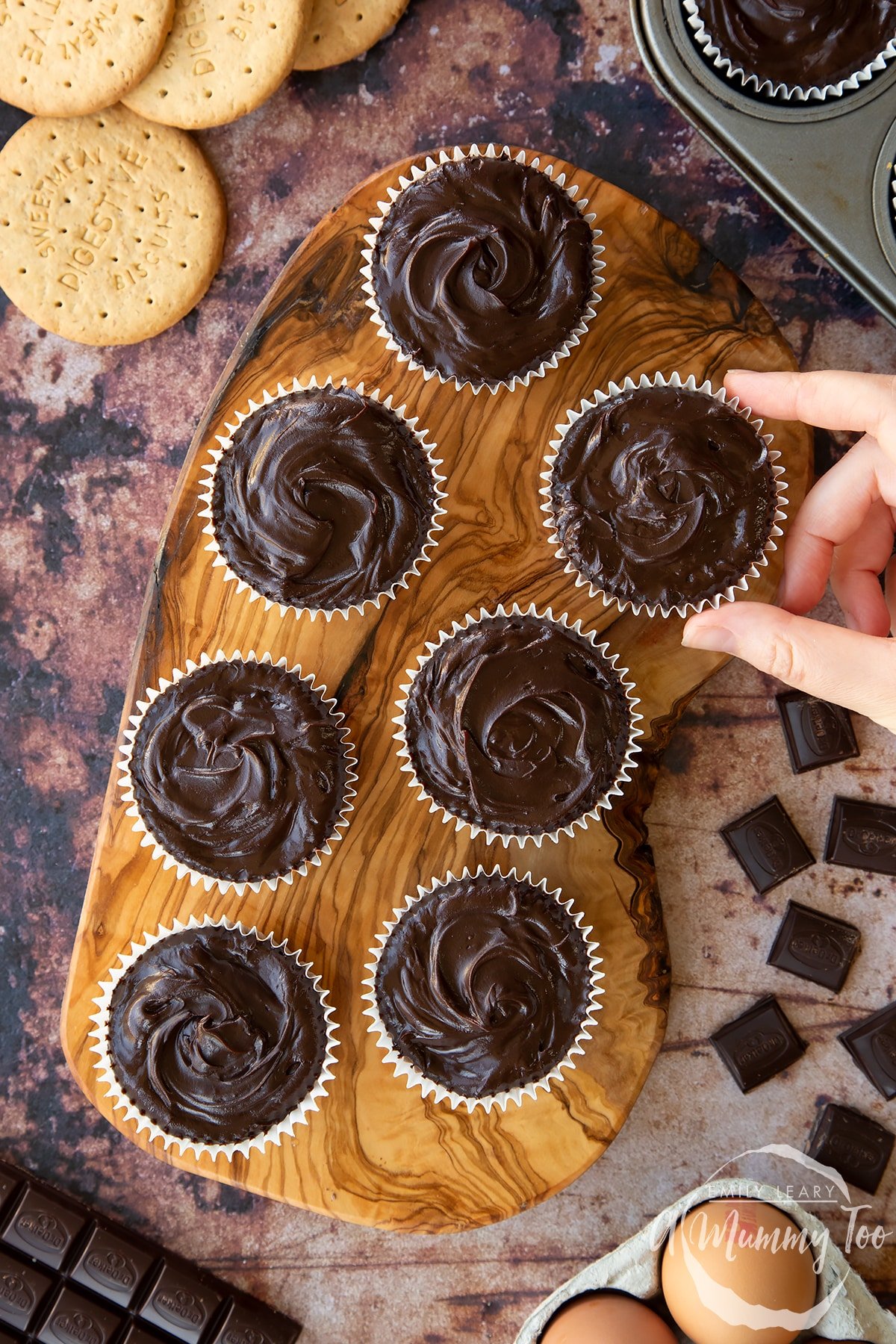 Chocolate cheesecake cupcakes in rows on an olive wood board, shown from above. A hand reaches for one.