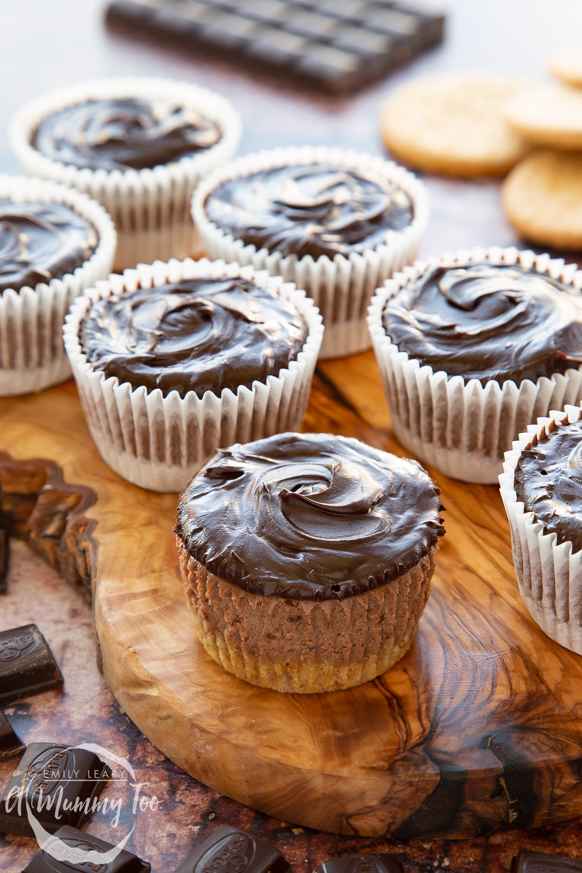 Chocolate cheesecake cupcakes in rows on an olive wood board. The one in the foreground has been unwrapped.