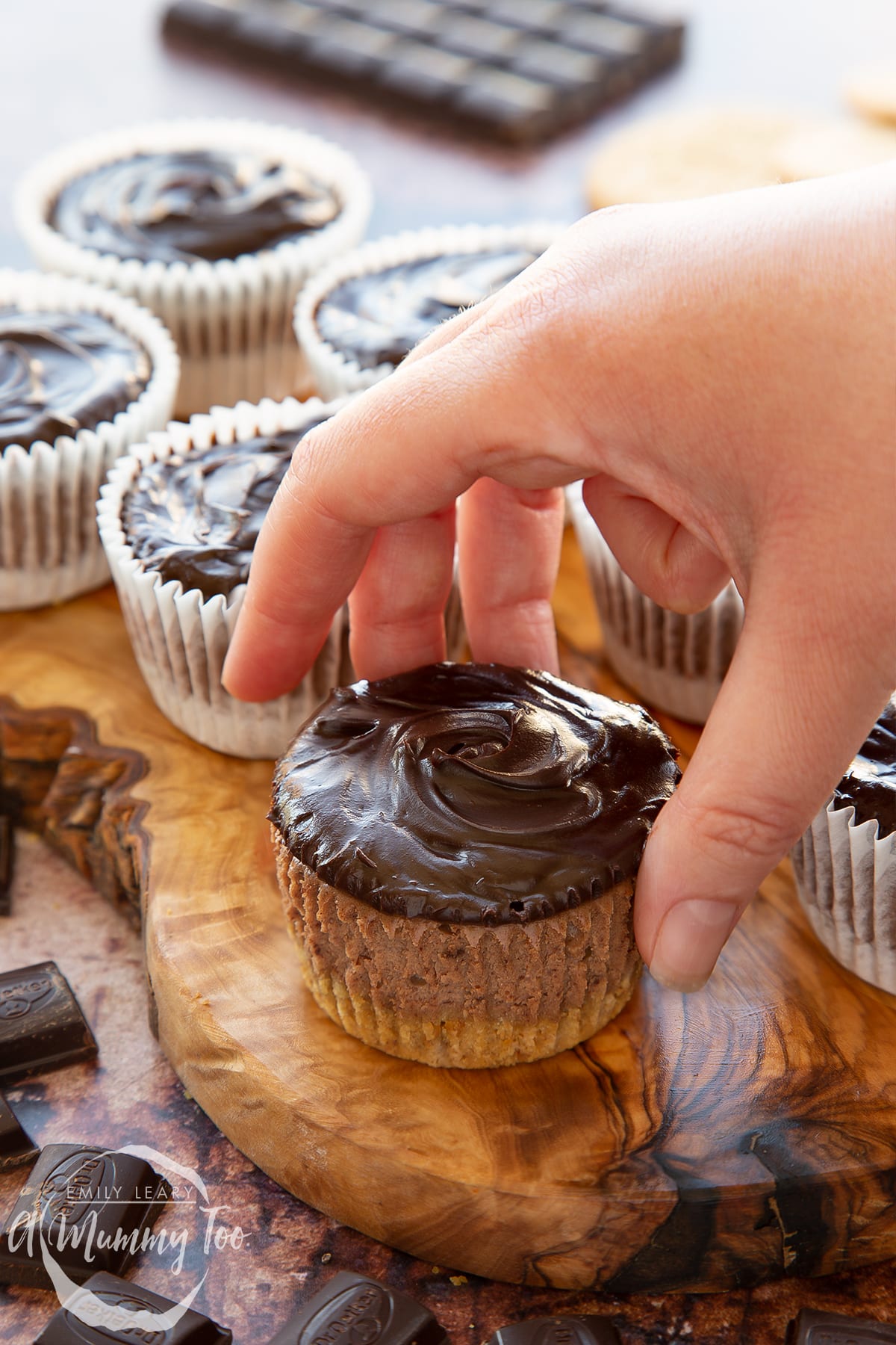 Chocolate cheesecake cupcakes in rows on an olive wood board. The one in the foreground has been unwrapped and a hand reaches for it.