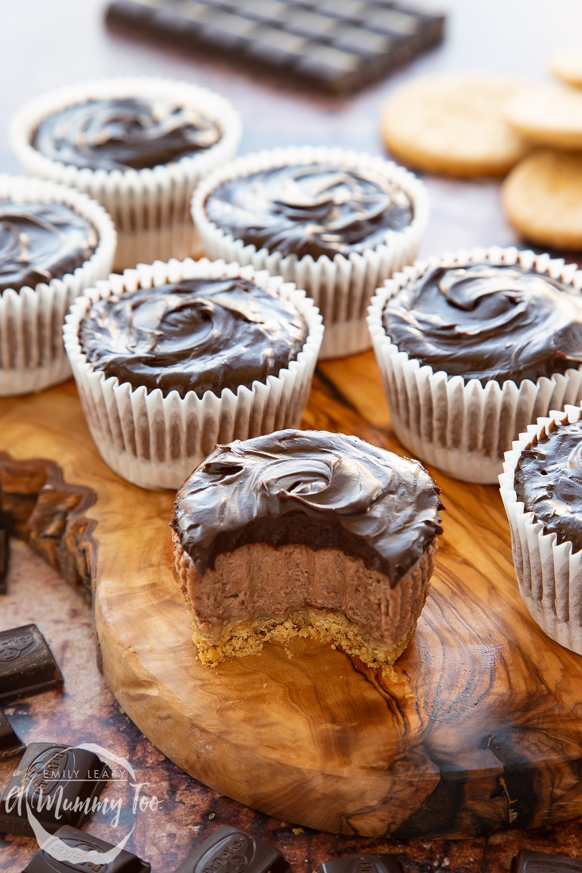 Chocolate cheesecake cupcakes in rows on an olive wood board. The one in the foreground has been unwrapped and has a bite out of it.