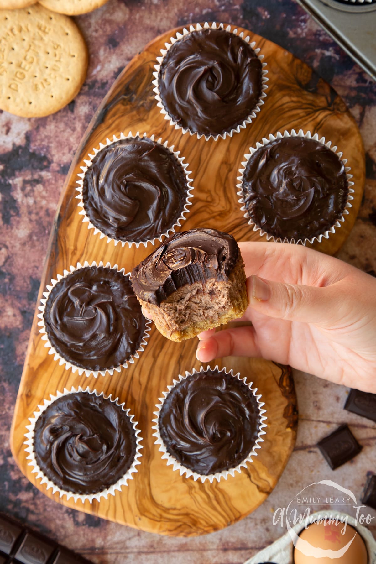 Chocolate cheesecake cupcakes in rows on an olive wood board, shown from above. A hand holds one with a bite out of it. 