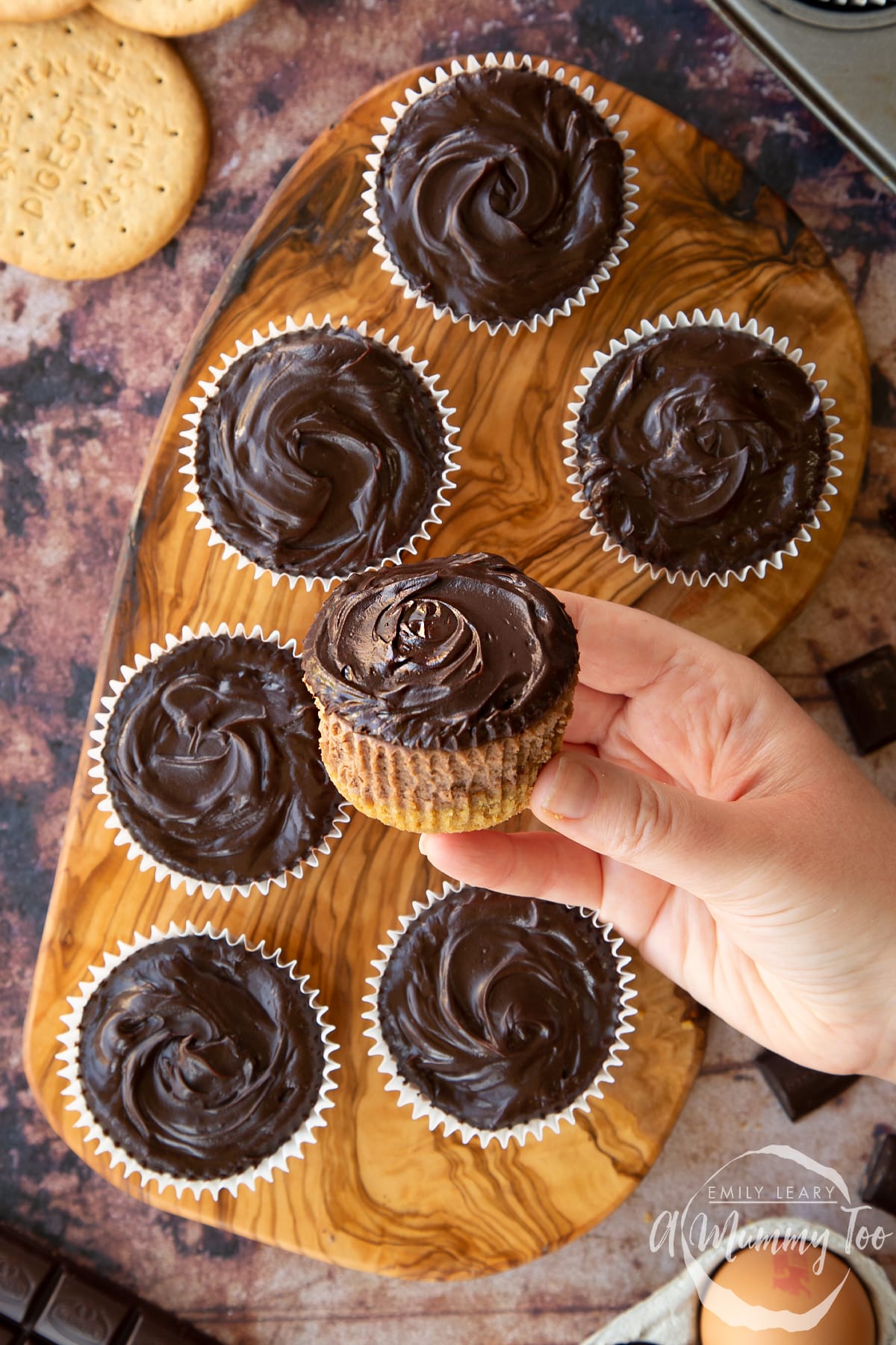 Chocolate cheesecake cupcakes in rows on an olive wood board, shown from above. A hand holds an unwrapped one.